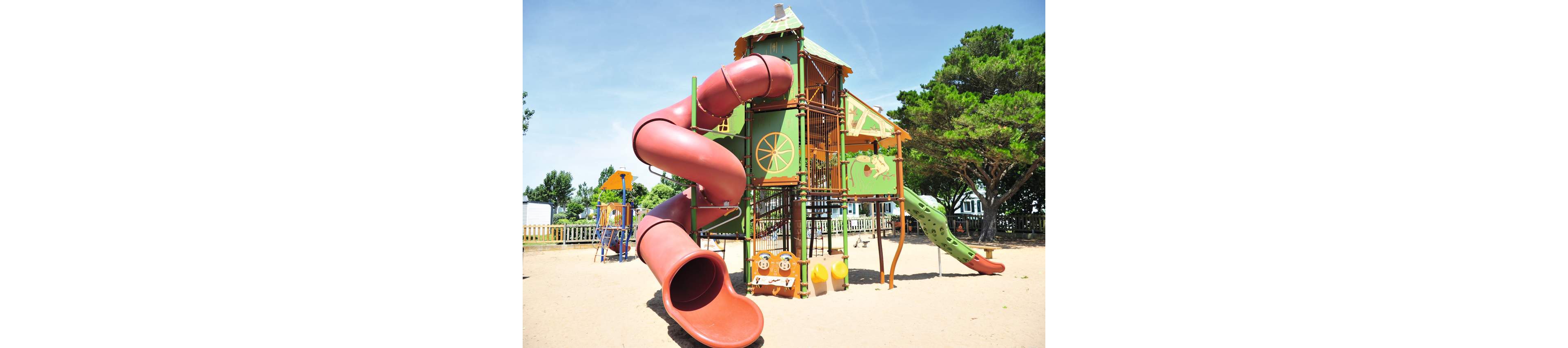 A large, colorful playground structure with multiple slides stands on a sandy surface. The main feature is a tall red spiral tube slide attached to a green and orange climbing tower. Additional play elements, including a green wavy slide and various climbing sections, extend from the structure. The playground is outdoors, surrounded by trees, fencing, and a clear blue sky, suggesting a bright summer day.