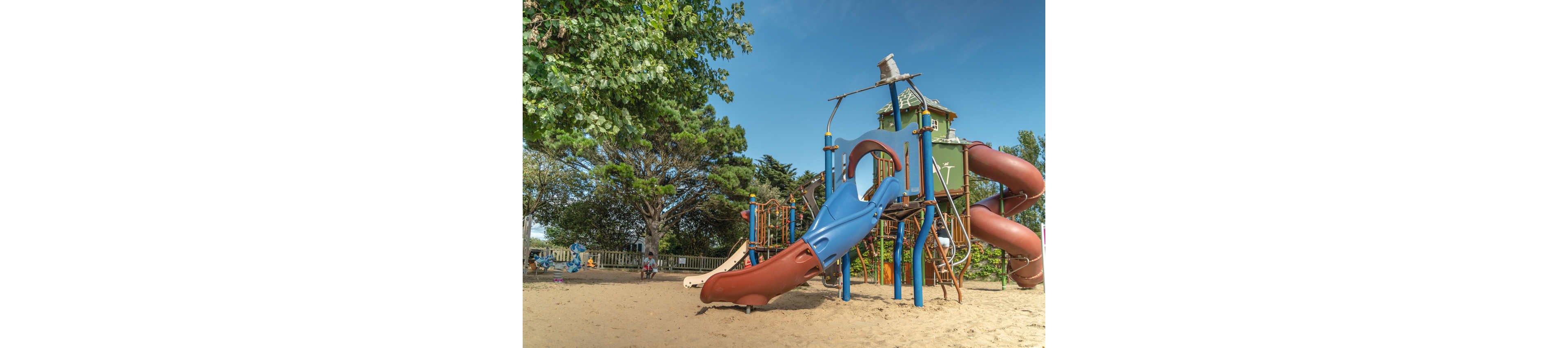A colorful playground structure with multiple slides stands on a sandy surface under a clear blue sky. The play area includes a large red spiral slide, a blue straight slide, and climbing elements connected to a tower-like structure. Tall green trees surround the playground, providing shade and a natural backdrop. A few children and adults are visible in the distance, enjoying the outdoor recreational space.