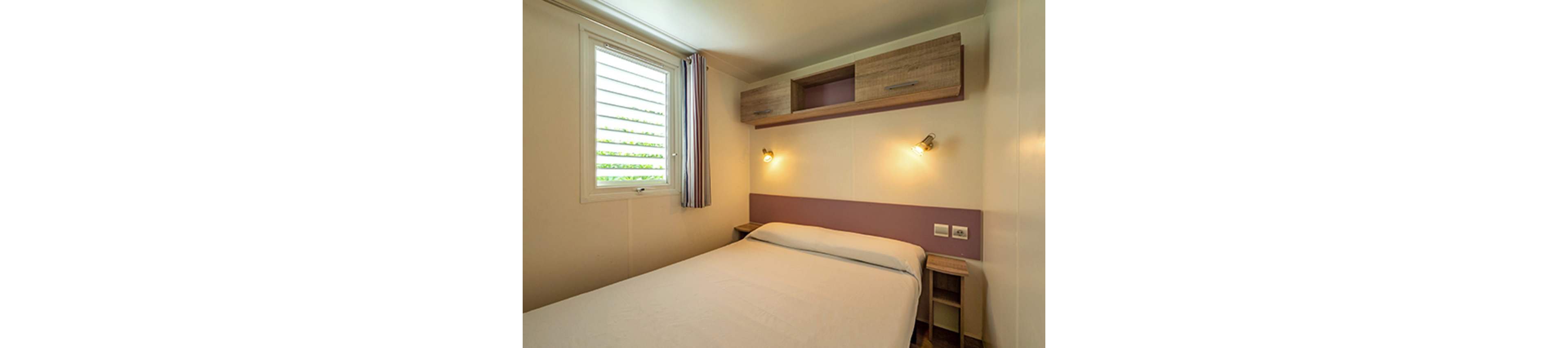 A small, cozy bedroom features a neatly made double bed with a white duvet. The room has light-colored walls with a mauve accent panel behind the bed. Two warm wall-mounted lamps provide soft lighting on either side of the headboard. A window with horizontal blinds and striped curtains lets in natural light, while wooden shelves and cabinets offer simple storage.