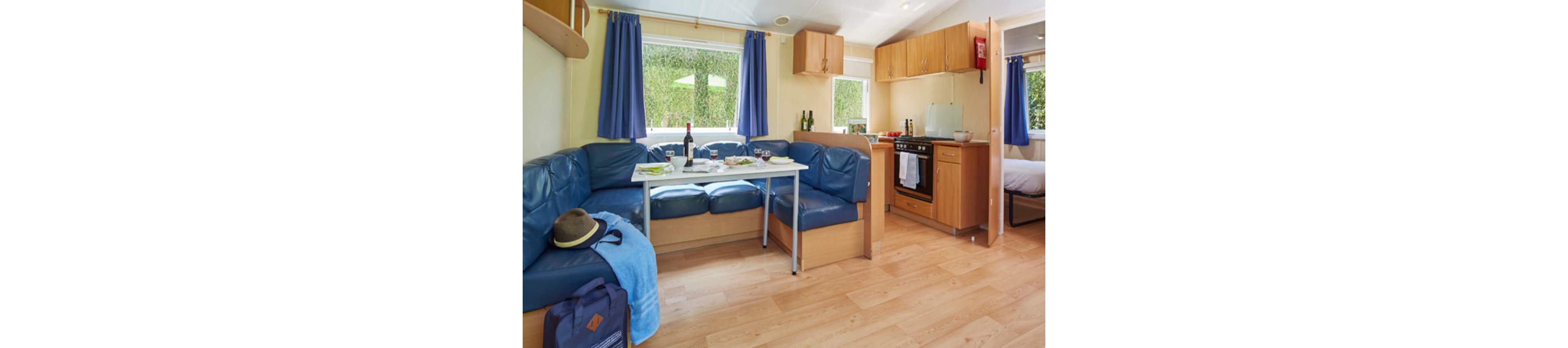Compact vacation accommodation interior featuring a small kitchen and dining area. A blue corner sofa surrounds a white dining table set with wine and snacks. Light wood cabinets, laminate flooring, and blue curtains create a bright, casual atmosphere. A backpack and hat on the seating suggest travel or a holiday stay. A partially visible bedroom and outdoor greenery through the windows complete the cozy setting.