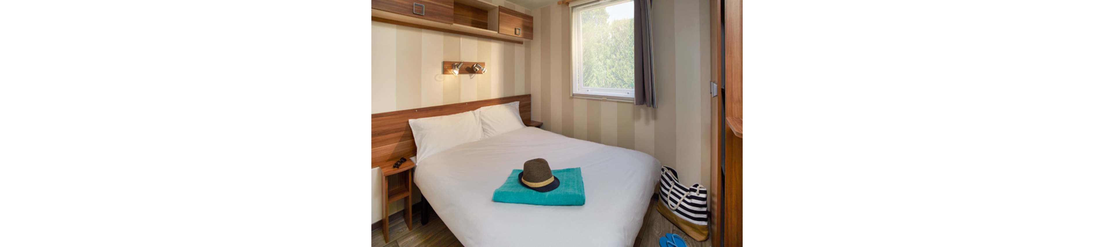 A small, cozy bedroom features a neatly made double bed with white linens. A folded turquoise towel and a straw hat are placed at the foot of the bed, suggesting a holiday or beach getaway. Wooden shelves and bedside tables frame the bed, while a window lets in natural light and shows greenery outside. A striped bag and blue sandals on the floor add to the relaxed vacation atmosphere.