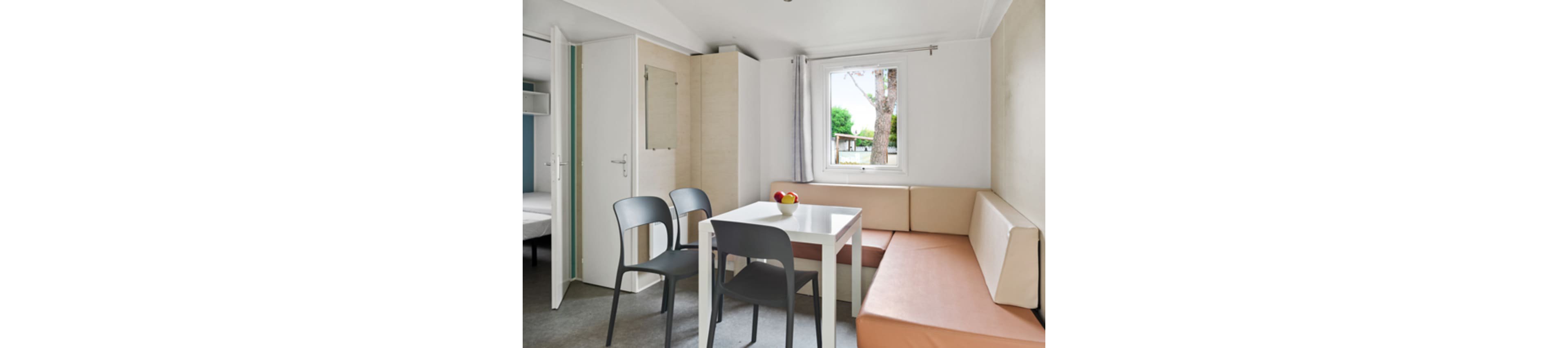 A compact dining area features a white table with three modern gray chairs and a built-in corner bench. The space is part of a small apartment or mobile home, with a bedroom visible through an open door. A window lets in natural light and offers a view of trees and greenery outside. Neutral tones and simple furnishings create a clean, minimalist atmosphere.