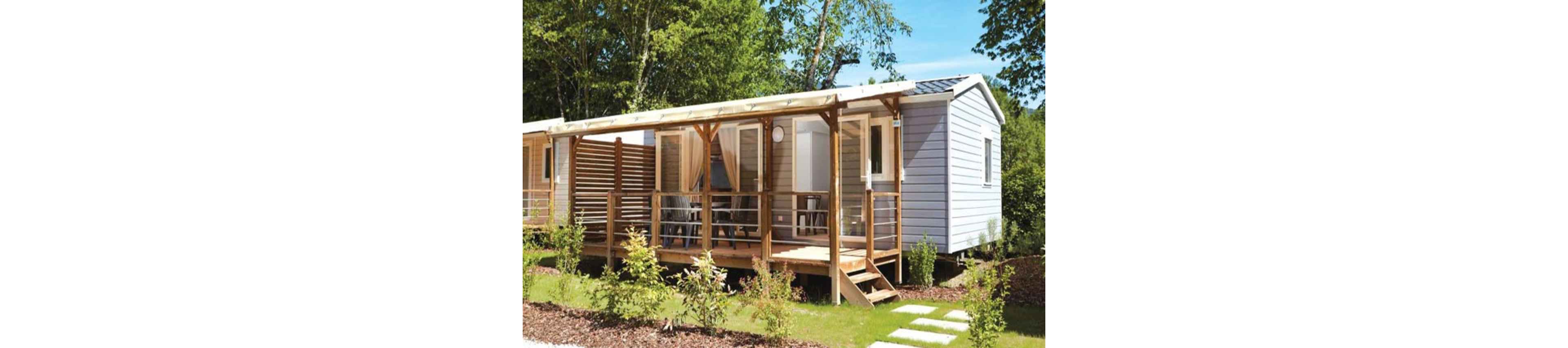 A modern mobile home with light-colored siding features a spacious wooden terrace and railing. Large windows and sheer curtains open onto the deck, creating an inviting indoor-outdoor living space. The home is set in a green, landscaped area with small shrubs, grass, and stepping stones. Tall trees in the background suggest a peaceful, natural holiday park or campsite setting.