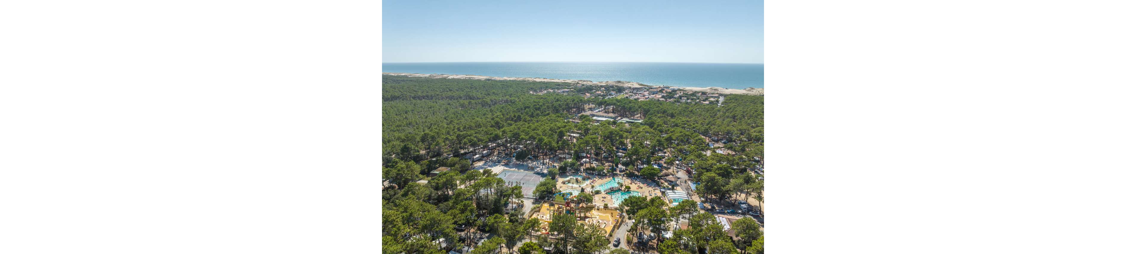 High above a dense coastal pine forest, a large holiday resort with pools and recreation areas is visible. The complex is nestled among the trees, with bright turquoise swimming pools standing out against the greenery. In the distance, a sandy shoreline and calm sea stretch across the horizon under a clear blue sky. The scene suggests a sunny, relaxed vacation destination surrounded by nature.