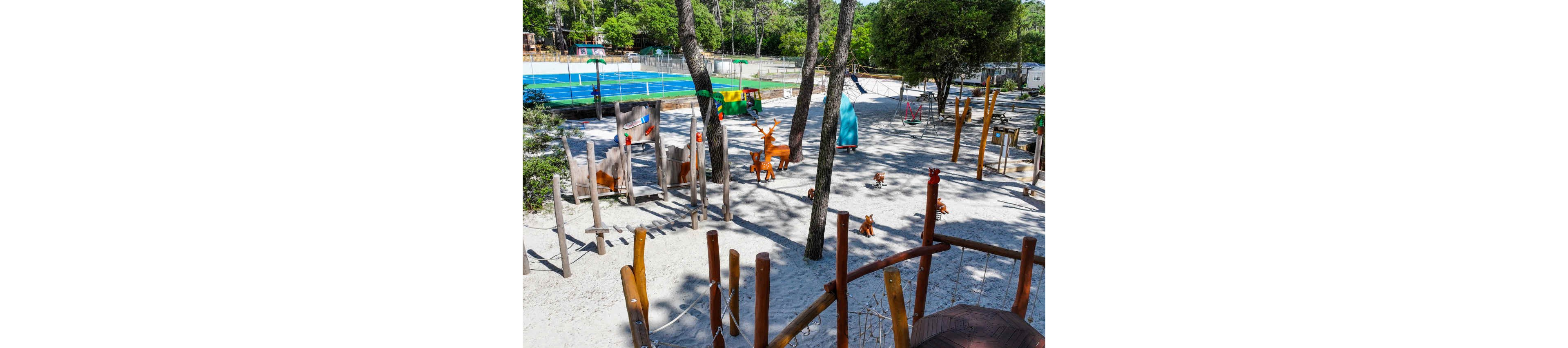 An outdoor playground is set in a sandy area surrounded by tall trees. Wooden climbing structures, balance beams, and rope elements are arranged across the play space. Colorful animal sculptures, including deer and foxes, are scattered around for children to explore. In the background, a bright blue sports court and lush green foliage add contrast to the sunny scene.