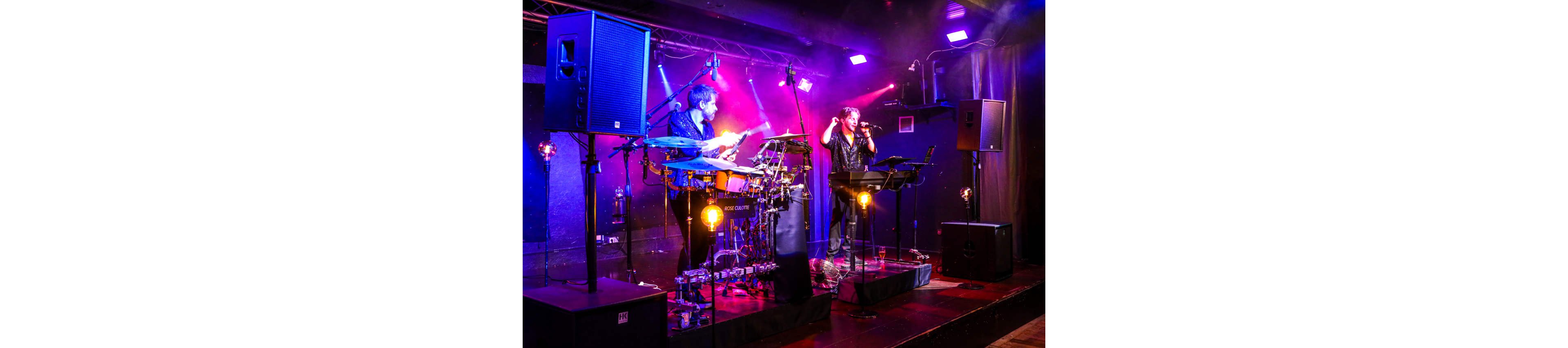 An electronic music duo performs on a small indoor stage surrounded by professional speakers and lighting. One musician plays an electronic drum kit while the other sings or speaks into a microphone behind a keyboard and control station. Vibrant purple, pink, and blue stage lights create a dramatic, smoky atmosphere around the performers. The scene suggests an intimate concert or club show focused on live electronic performance.