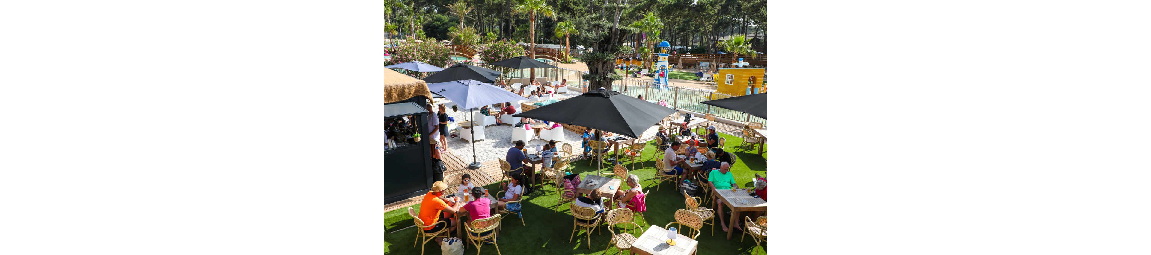 An outdoor café terrace is filled with people sitting at tables under large umbrellas. Families and friends enjoy food and drinks on a sunny day, surrounded by greenery and palm trees. In the background, a fenced water play area with slides and colorful structures entertains children. The scene conveys a relaxed holiday atmosphere at a leisure resort.