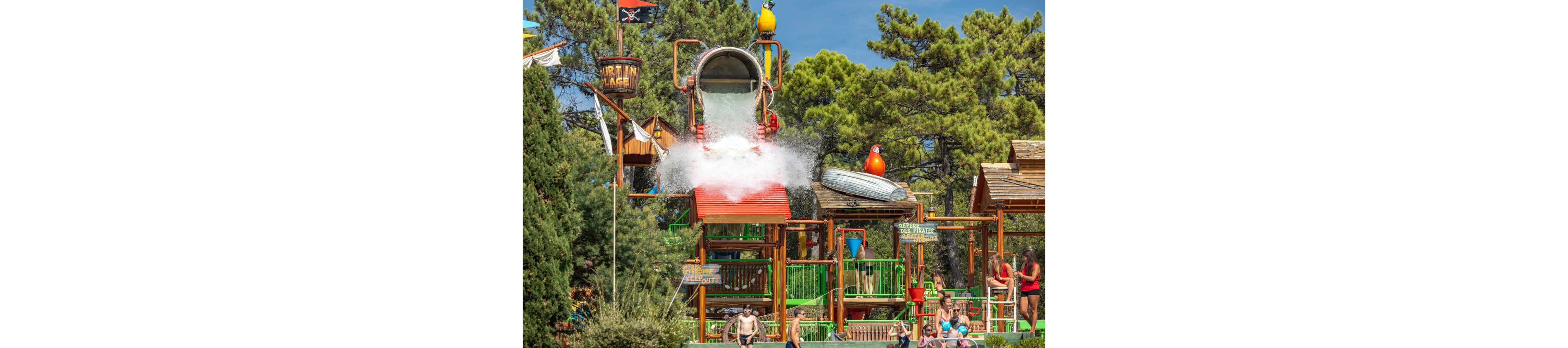 A giant tipping bucket dumps a huge splash of water onto a colorful multi-level water play structure. The scene is set in an outdoor water park surrounded by tall green trees. Children and adults stand and walk around the attraction, some waiting for the next splash while others watch from nearby. Bright slides, ladders, and playful decorations create a lively, fun summertime atmosphere.