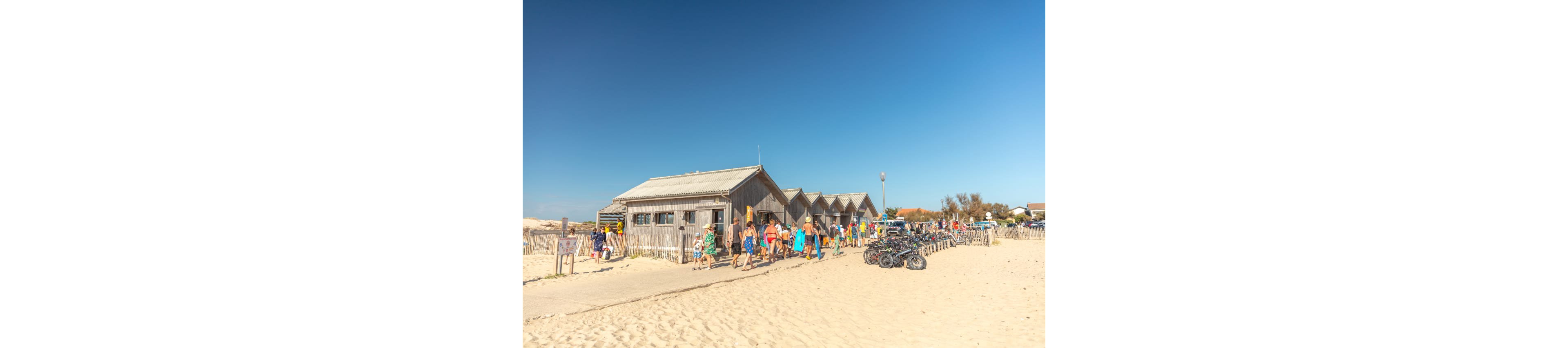 A group of people walk along a sandy path beside a row of wooden cabins near the beach. The bright blue sky and strong sunlight suggest a warm summer day. Bicycles are parked in the sand near a fence, indicating visitors arriving by bike. The scene captures a relaxed holiday atmosphere at a coastal destination.