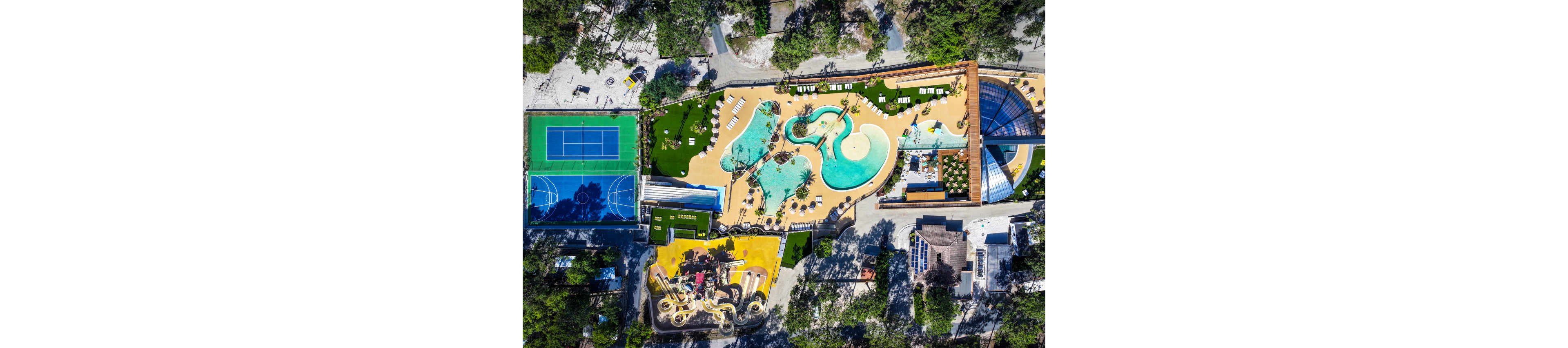 A colorful resort complex is captured from above, featuring an expansive freeform swimming pool surrounded by lounge chairs and umbrellas. Adjacent to the pool area are twisting water slides and a separate splash zone. A full-size tennis court and a multi-use sports court sit nearby, bordered by trees and landscaped paths. The scene suggests a lively vacation destination with multiple outdoor recreation options.