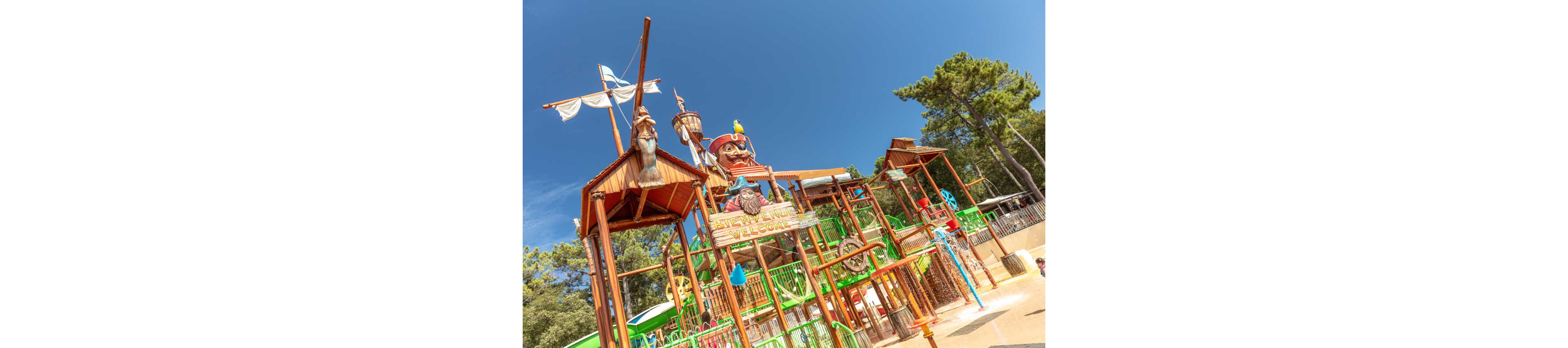 A vibrant pirate-themed water play structure stands under a clear blue sky. The scene features wooden towers, slides, and climbing elements decorated with pirate characters and nautical details. Bright green, orange, and blue accents create a playful, family-friendly atmosphere. Tall trees surround the attraction, emphasizing its outdoor amusement park setting.