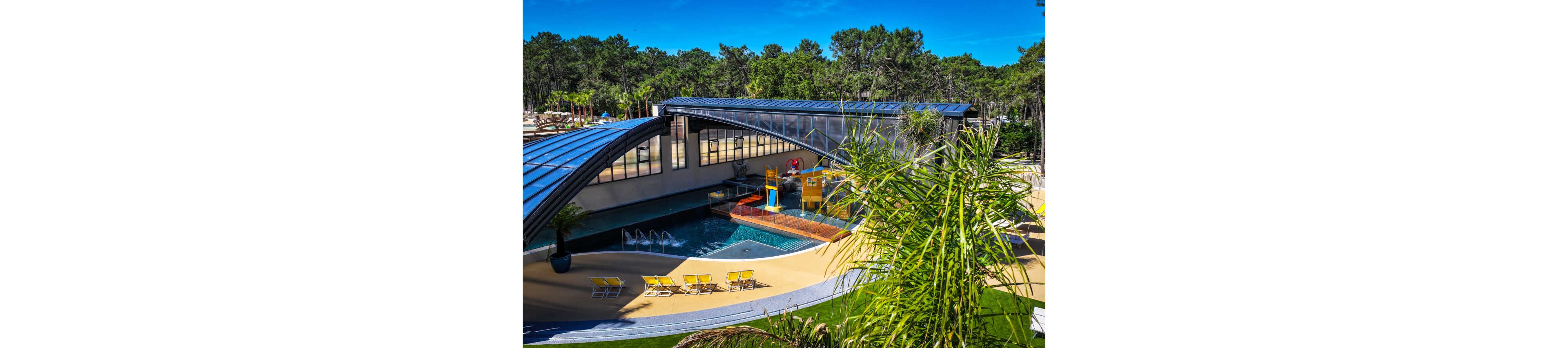 A contemporary swimming pool complex sits nestled among tall pine trees at a resort. The main pool area features a partially covered structure with large glass panels and a curved metal roof. Bright yellow lounge chairs and a small water slide add playful color to the scene. Lush greenery and clear blue skies create a relaxing holiday atmosphere.