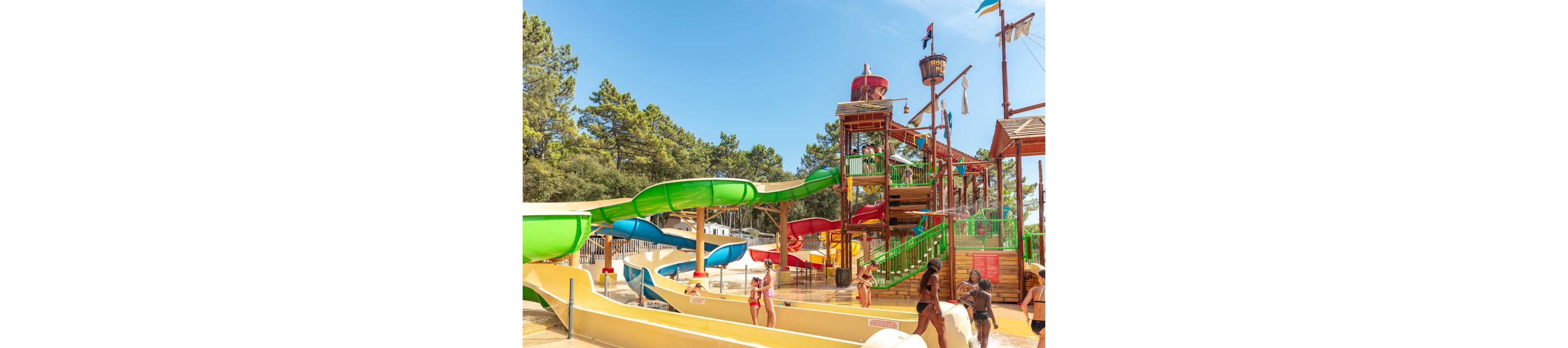 An outdoor water park features multiple colorful slides and a large play structure with stairs, platforms, and water features. Several people in swimsuits, including adults and children, walk and play around the shallow splash area at the slide exits. Tall green trees surround the park under a bright blue sky, creating a sunny summer atmosphere. The scene conveys leisure, fun, and family-friendly recreation in a natural setting.