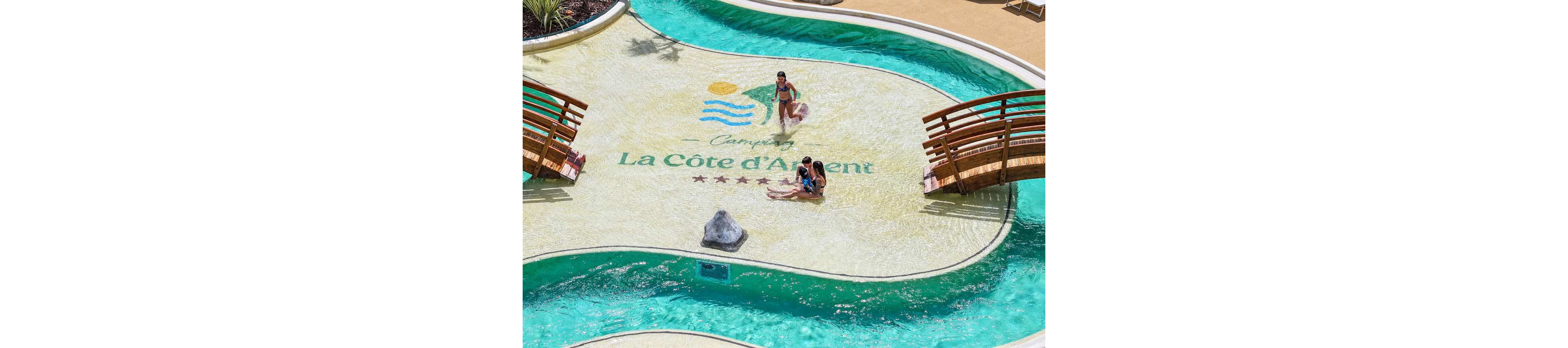 A shallow resort pool features a central sand-colored island with the text "Camping La Côte d’Argent" and four stars. Two small wooden bridges curve over the surrounding turquoise water. An adult and child sit together at the water’s edge while another adult stands nearby, all dressed in swimwear. The scene suggests a relaxed family holiday atmosphere at a sunny campground resort.