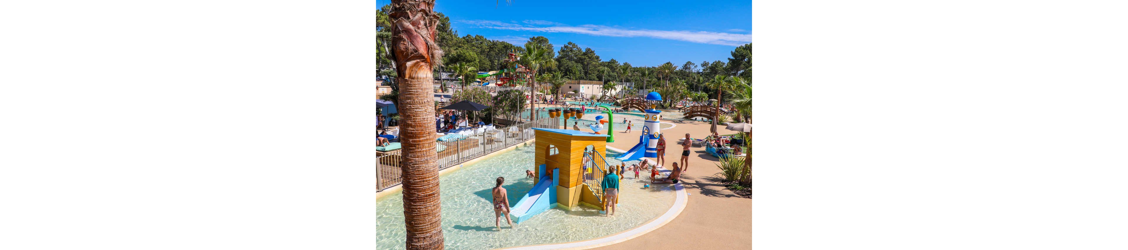 An outdoor water playground features shallow pools with small slides and brightly colored play structures. Families with children splash and relax under the sun, surrounded by palm trees and lounge chairs. The scene includes a lighthouse-themed structure, a wooden ship play area, and multiple water features. Clear blue skies and lush greenery create a vibrant, summery holiday atmosphere.