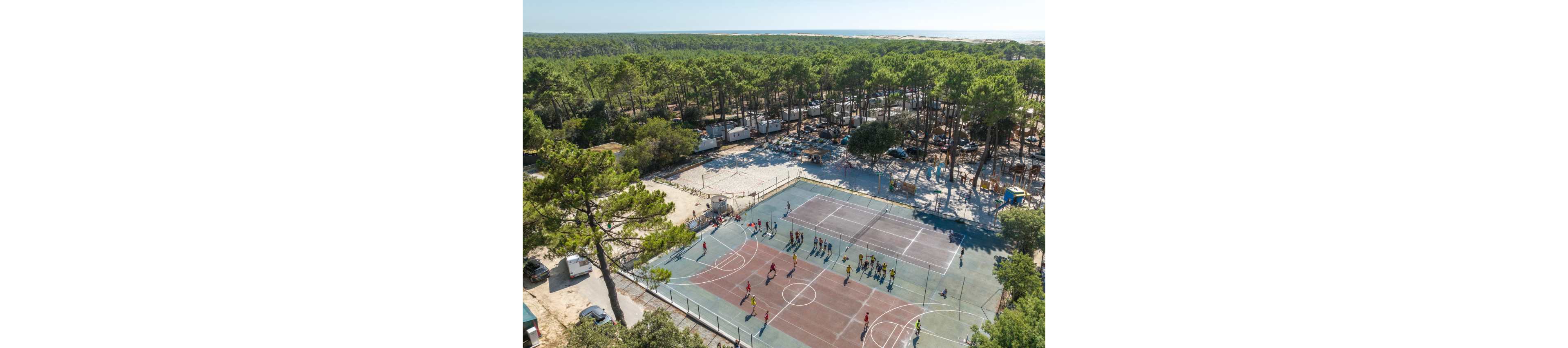 A large outdoor sports court is captured from above, surrounded by tall pine trees and a busy campsite. Groups of people play games on the multi-colored court while others stand along the sidelines. Nearby, tents, caravans, and vehicles are scattered among the trees, suggesting a holiday or summer camp atmosphere. In the distance, the forest stretches toward a bright horizon, giving the scene an open, recreational feel.