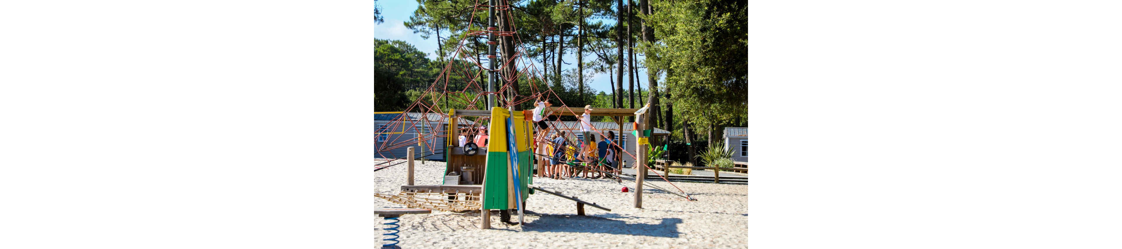 A group of children play together on a large rope climbing structure in a sandy outdoor playground. The play area features wooden equipment painted in bright colors and surrounded by tall pine trees. Some kids climb the ropes while others gather underneath and around the structure. In the background, small cabins or mobile homes suggest a campsite or holiday park setting.
