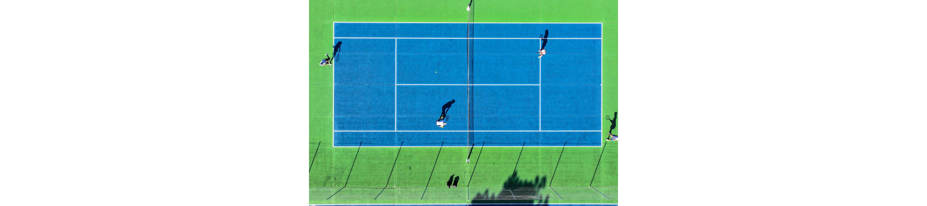 Overhead view of a bright blue tennis court surrounded by green surface. Four players are positioned on opposite sides of the net, actively engaged in a match. Long shadows stretch across the court, suggesting strong sunlight and a clear day. The geometric lines of the court create a graphic, high-contrast composition.