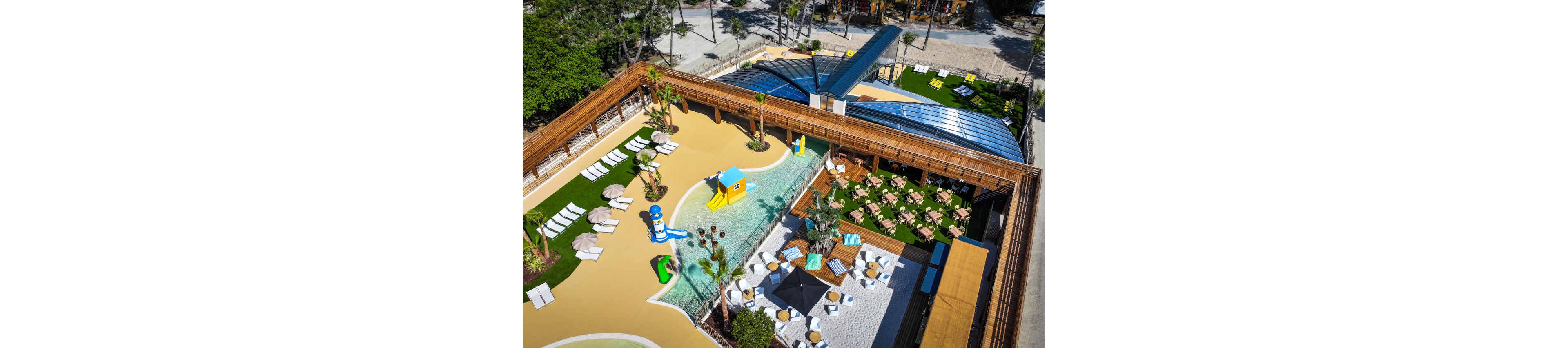 A contemporary outdoor resort area is captured from above, featuring a shallow pool with colorful slides and a small play structure. Surrounding the water are sun loungers, umbrellas, and shaded seating areas arranged on artificial grass and sand-colored surfaces. A large wooden deck and covered pavilion frame the pool complex, creating distinct zones for relaxation and recreation. The scene is bright and sunny, emphasizing the clean lines, vibrant colors, and family-friendly design of the leisure space.