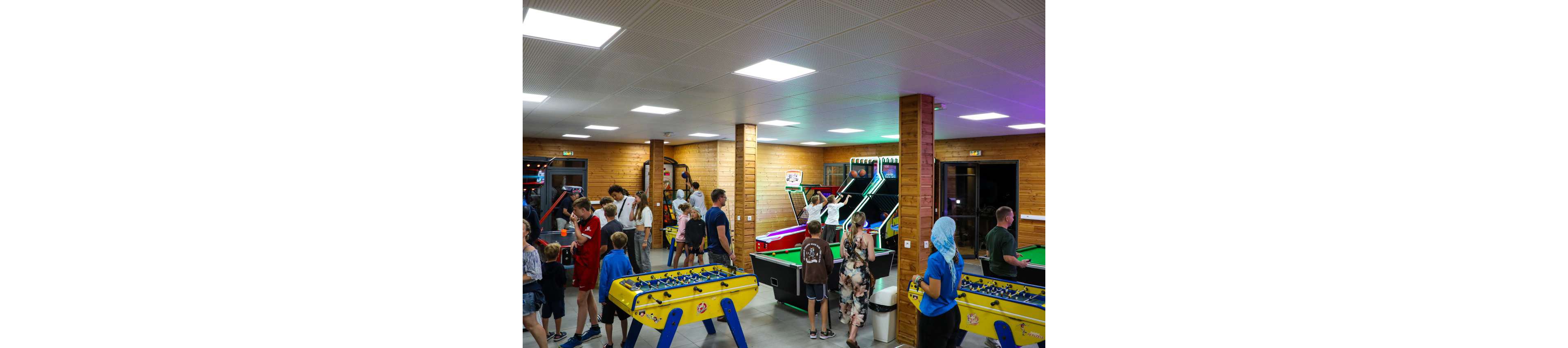 A busy indoor arcade is filled with people playing various games. Children and adults gather around foosball tables, arcade basketball machines, and pool tables. The wooden walls and bright ceiling lights create a warm, casual atmosphere. Players are actively engaged, chatting and moving between different game stations throughout the room.
