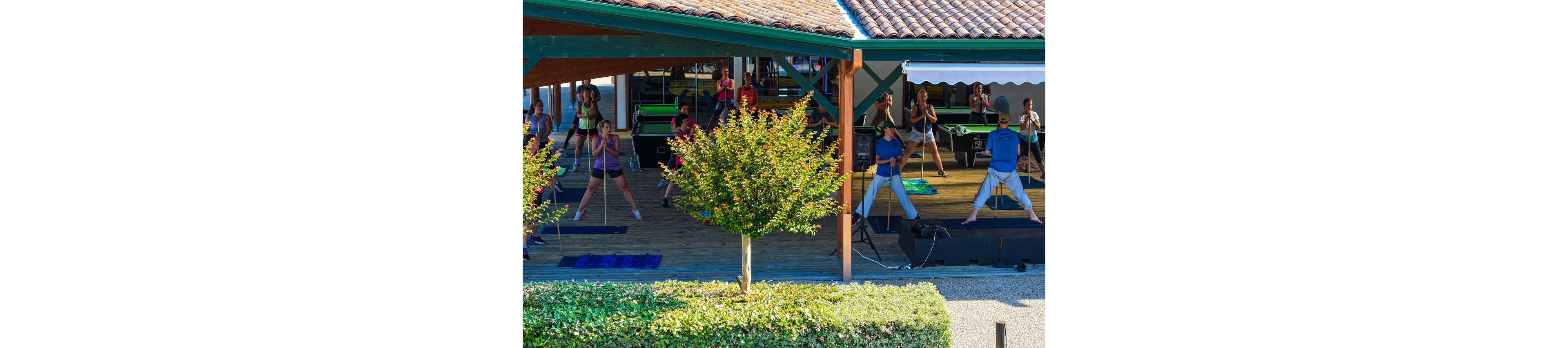 A large group of adults participates in an outdoor fitness class under a covered pavilion. The instructor stands on a small stage at the front, leading stretching or aerobic exercises with long sticks. Participants are spaced out on exercise mats, following the movements in unison. Greenery and small trees frame the scene, creating a bright and energetic atmosphere in a recreational setting.