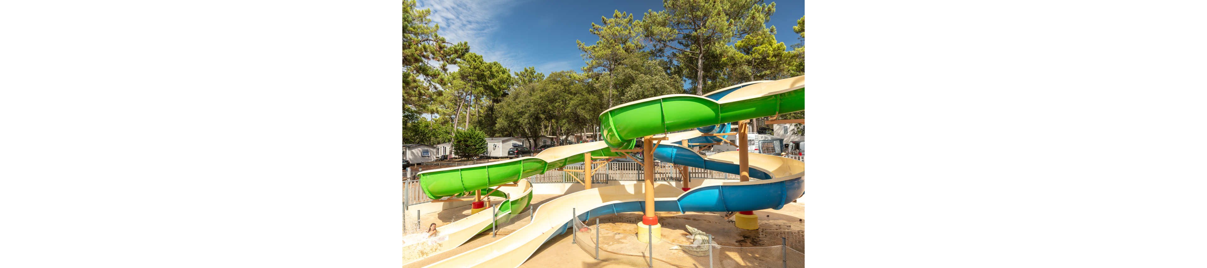 A large outdoor waterslide with green and blue enclosed tubes twists above a sandy ground. The slide ends in a shallow splash area where a child is visible enjoying the ride. Tall pine trees and several camper vans or mobile homes surround the water attraction, suggesting a holiday campground setting. The bright daylight and clear blue sky create a cheerful, summery atmosphere.