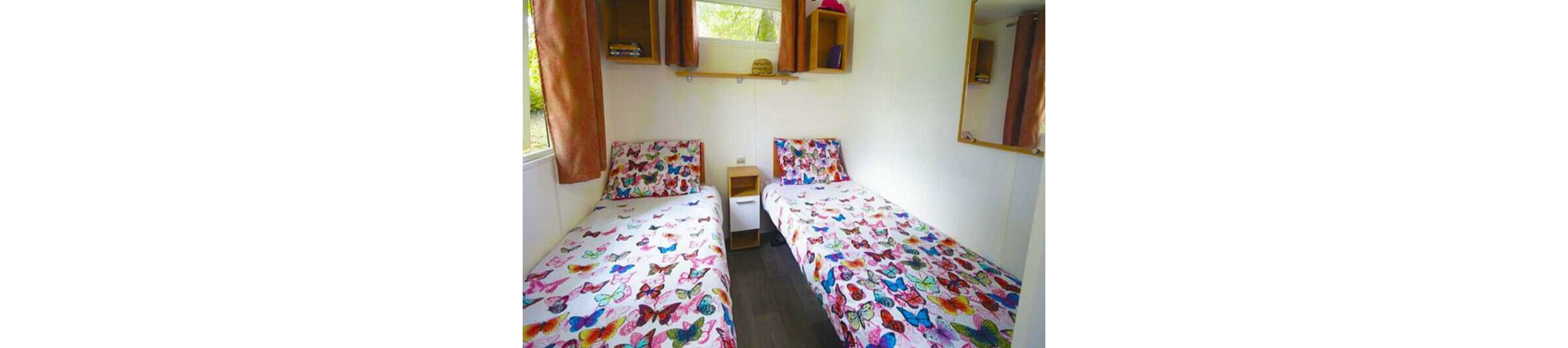 A compact bedroom features two single beds with colorful butterfly-patterned bedspreads. The beds are separated by a small white nightstand against a light-colored wall. Wall-mounted shelves with books and decor, along with a mirror, add storage and visual interest. Natural light enters through a window with curtains, creating a cheerful and cozy atmosphere.