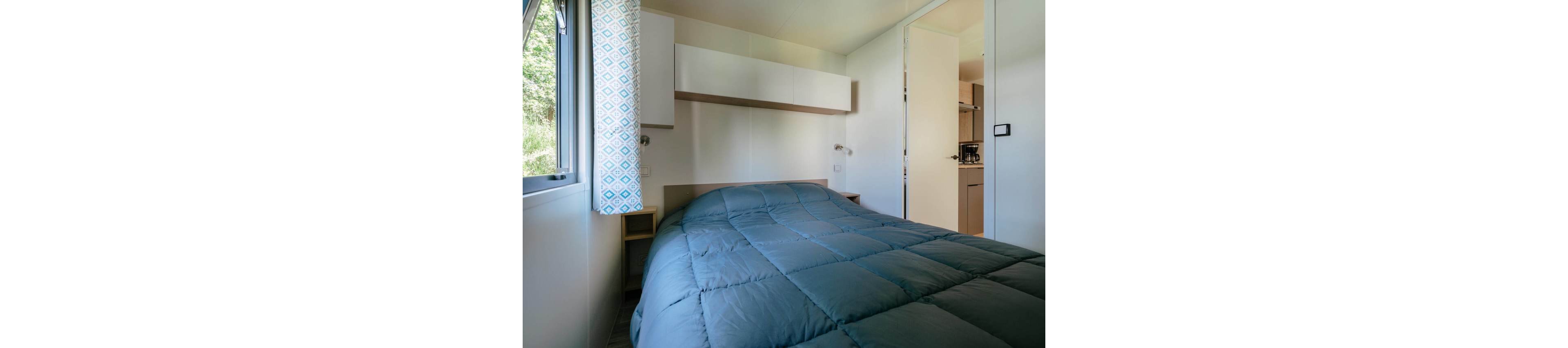 A small bedroom features a neatly made bed with a teal quilted comforter. Natural light enters through a window with patterned curtains on the left side. Minimalist white walls and overhead storage cabinets create a clean, modern look. An open door reveals a glimpse of an adjacent kitchenette area, emphasizing the compact living space.