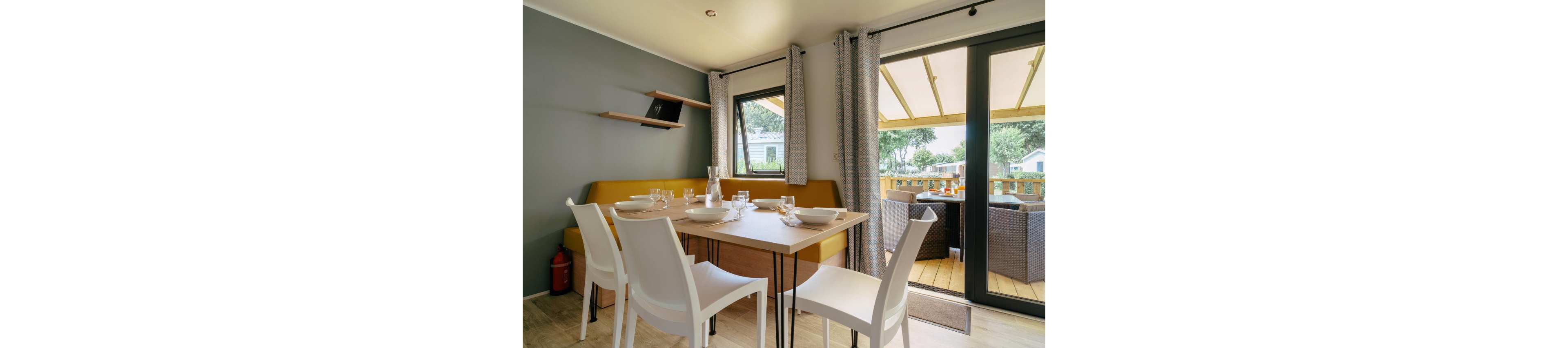 Modern dining space with a wooden table and white chairs is set with plates, glasses, and a carafe. A built-in yellow bench lines the wall beneath a small floating shelf. Large windows and glass doors with patterned curtains open onto a covered wooden terrace with wicker-style outdoor seating. Natural light and neutral tones create a cozy, inviting atmosphere suitable for vacation or small family meals.