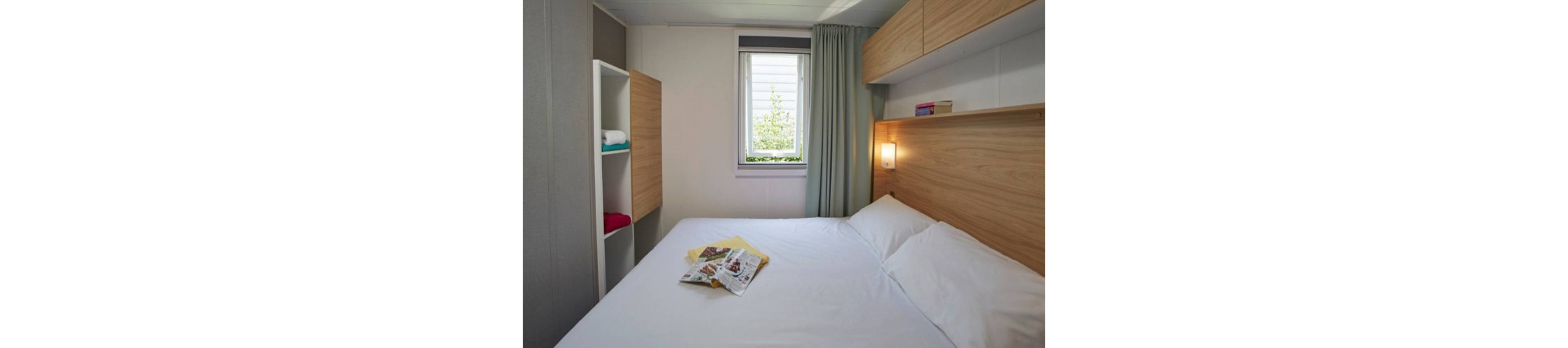 Compact modern bedroom featuring a neatly made double bed with white linens. Light wood cabinets and shelving provide storage along the wall and beside the bed. A small window with curtains lets in natural light, illuminating a wall-mounted bedside lamp. Magazines and folded towels rest on the bed, adding a lived-in yet tidy feel to the space.