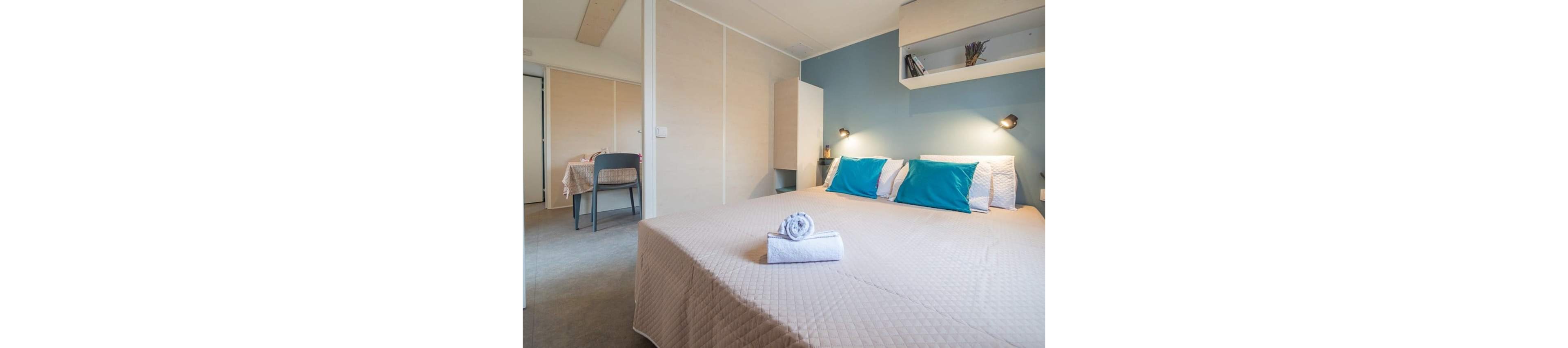 A neatly made double bed with teal and white pillows is the focal point of this modern bedroom. Soft neutral walls and a blue accent wall create a calm, contemporary atmosphere. Folded towels are placed on the bed, suggesting a guest-ready space. Built-in shelves, bedside lamps, and a small adjoining room with a table and chair add functional comfort to the interior.