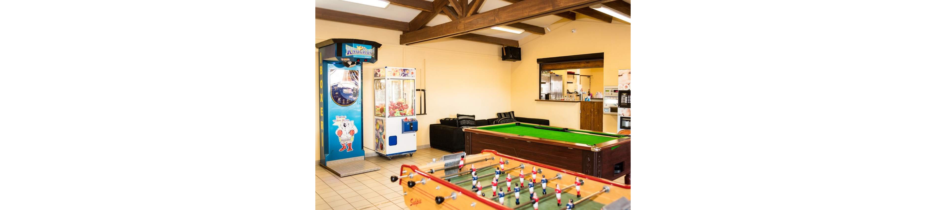 A bright recreation room features a foosball table in the foreground and a green-felt pool table behind it. On the left side, an arcade machine and a claw crane game stand against the wall. In the background, a black sofa seating area faces the games, and a service counter or kitchenette is visible. Exposed wooden beams and warm lighting create a casual, welcoming atmosphere suitable for leisure and socializing.