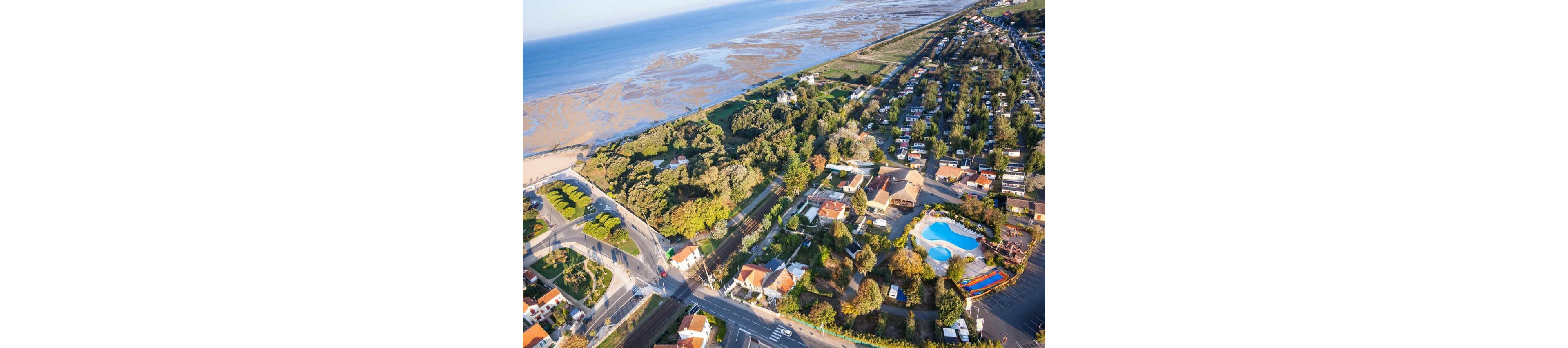 An aerial view captures a coastal residential neighborhood beside a shoreline. Houses with red and light-colored roofs are arranged along tree-lined streets and small roads. A bright blue swimming pool and recreational area stand out among the buildings. The coastline and tidal flats stretch into the distance, contrasting with the green vegetation and urban layout.