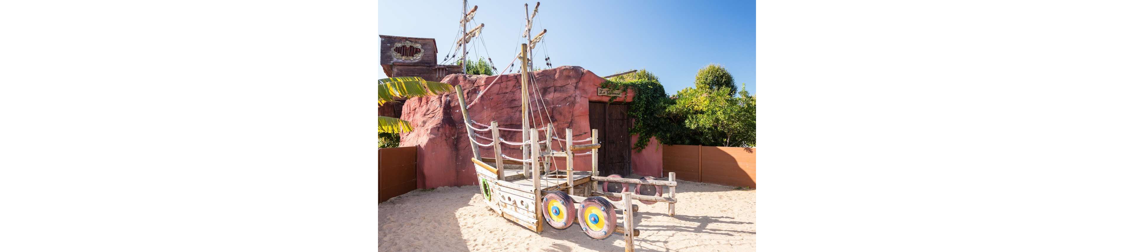 A small wooden pirate ship playground structure stands on a sandy surface under a clear blue sky. The play ship features colorful circular shields on its side and simple rope railings. Behind it, a rock-like red wall and wooden structures suggest a themed adventure setting. Green foliage and brown fencing frame the scene, creating a bright and playful outdoor environment.