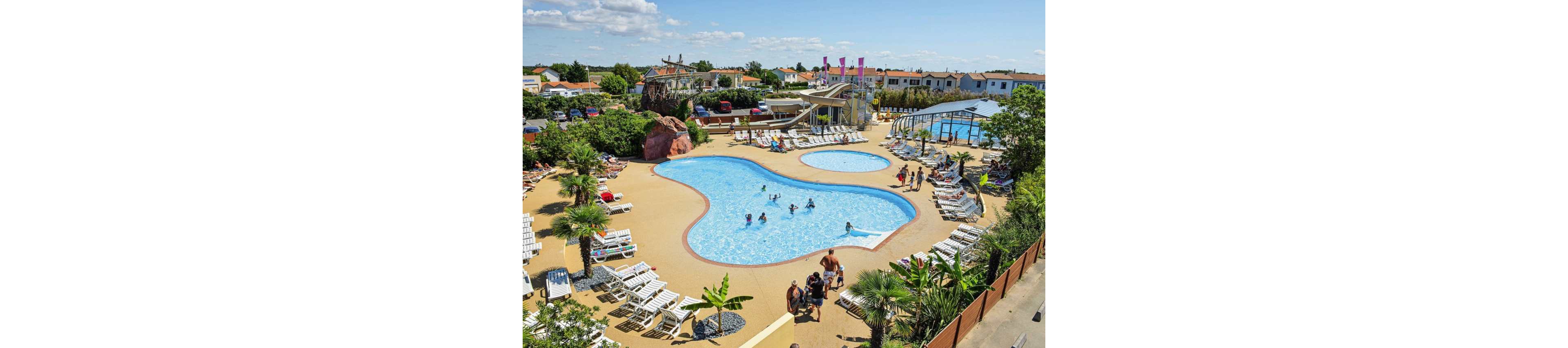 An outdoor resort features a large freeform swimming pool surrounded by sun loungers and palm trees. Families and children enjoy the water while others relax on deck chairs under the bright sun. A waterslide structure and additional smaller pools add to the recreational area. In the background, low-rise holiday homes and a covered pool are visible under a blue sky with scattered clouds.