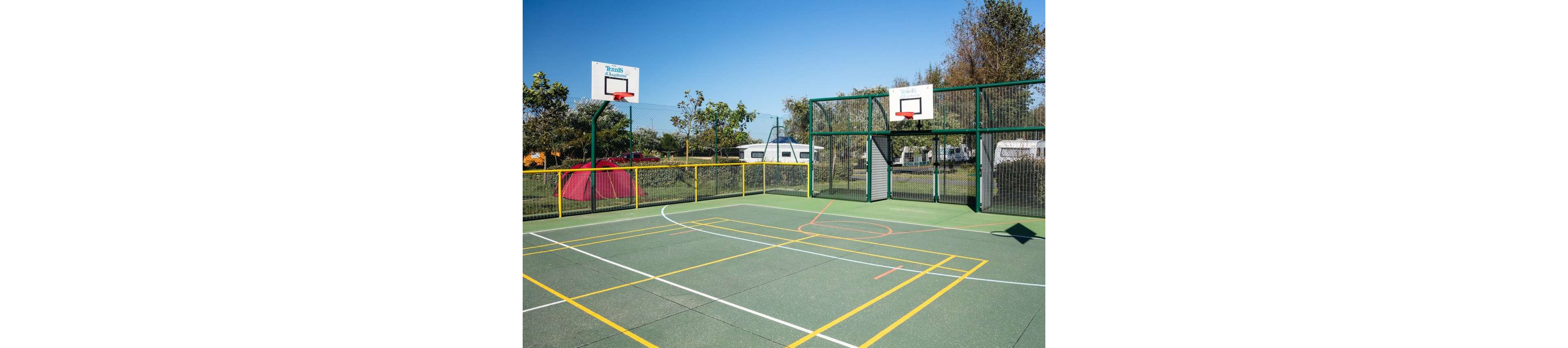 An empty outdoor multi-sport court features two basketball hoops at opposite ends. The green playing surface is marked with yellow and white lines for different sports. A fenced perimeter surrounds the court, with trees, grass, and parked caravans visible in the background. A red tent stands near the fence, suggesting a campsite or recreational park setting. Bright daylight and clear skies create a clean, inviting atmosphere for sports and leisure.