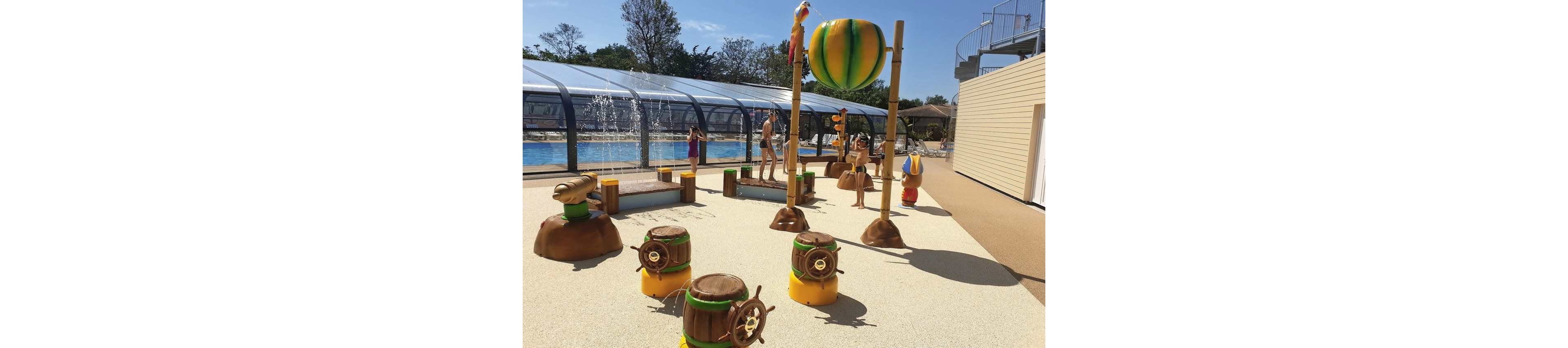 An outdoor splash play area features colorful cartoon-style structures near a swimming pool. Children play among barrel-shaped fountains, character statues, and a large suspended water bucket. A covered pool with water jets is visible in the background, along with a spiral staircase and sunlit deck. The scene suggests a family-friendly water park on a bright, sunny day.