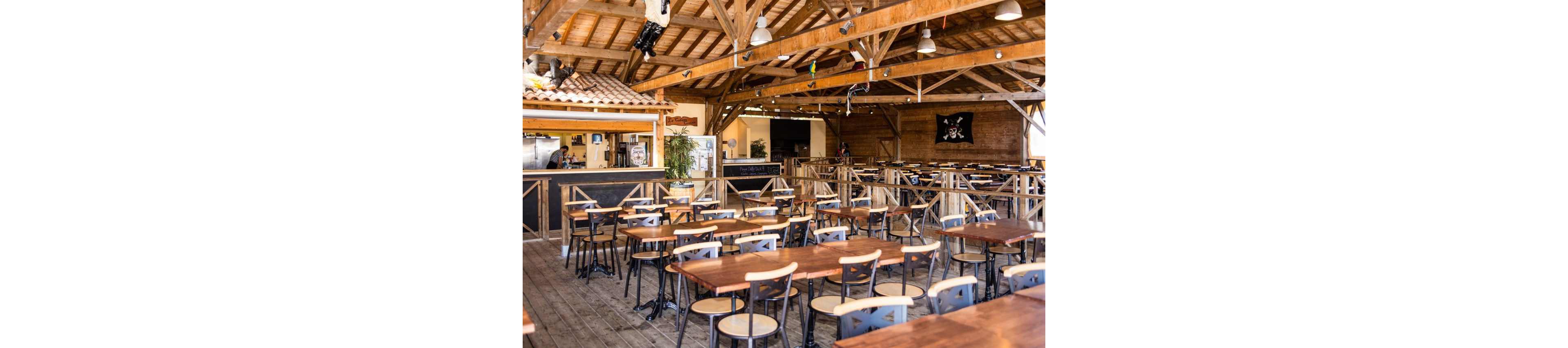 A spacious open-air restaurant features rows of wooden tables and metal-framed chairs under a high timber roof. The interior has a rustic, beach-bar feel with exposed beams, string lights, and a small bar area in the background. A pirate-themed flag and decorative elements hang on the walls, adding a playful touch. The space appears ready for guests, with neatly arranged seating and warm natural lighting.