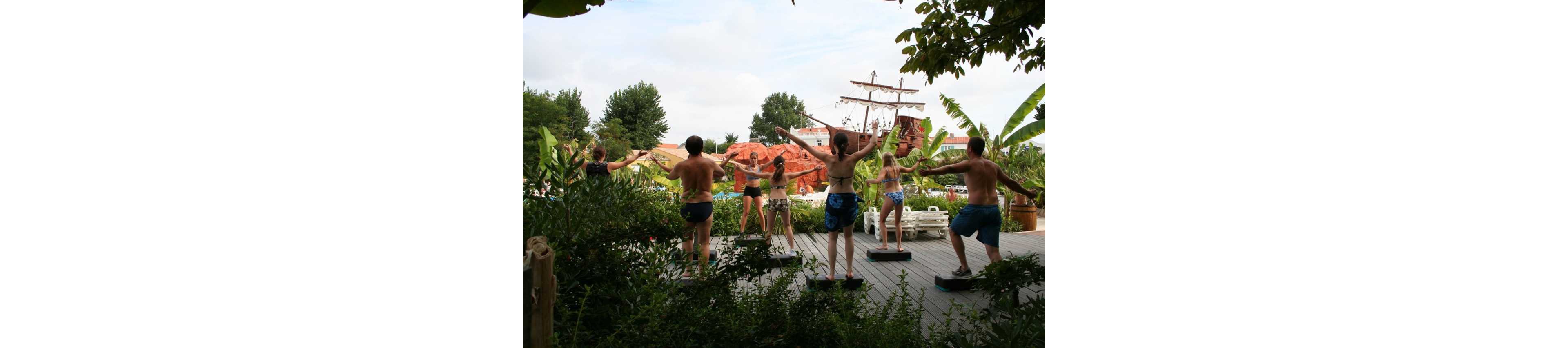 A group of people in swimwear participate in an outdoor fitness or exercise class. They stand on individual platforms on a wooden deck surrounded by lush tropical plants. In the background, a large decorative pirate ship and themed structures suggest a resort or water park setting. The scene feels casual and energetic, with participants stretching their arms in unison.