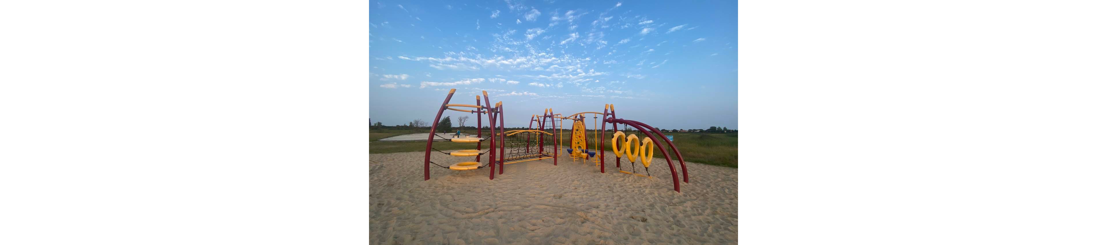 A colorful modern playground climbing structure stands on a sandy surface in an open outdoor area. The equipment features red frames with yellow climbing elements, rings, and nets designed for children’s play. The playground is surrounded by grassy fields under a wide blue sky with scattered clouds. No people are present, giving the scene a calm and quiet atmosphere.