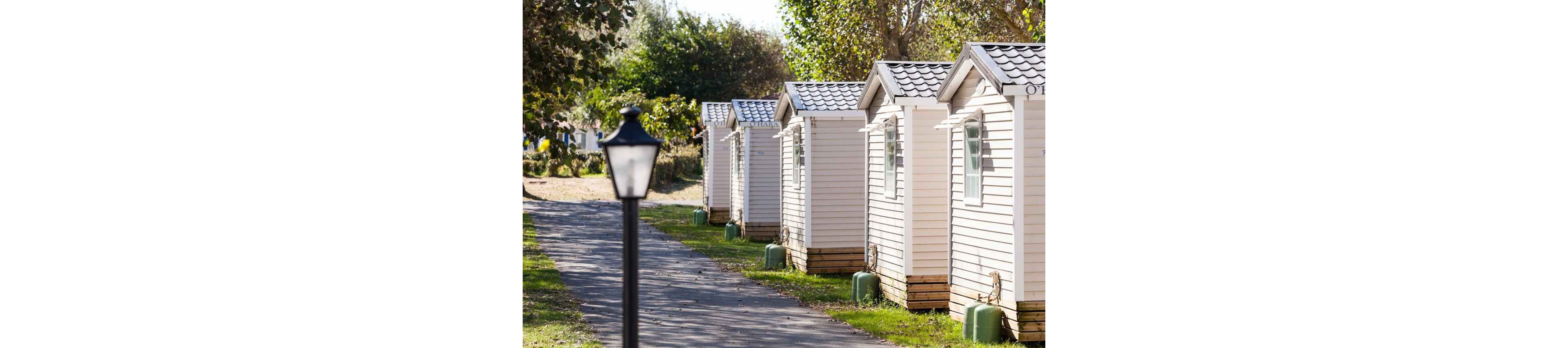 A row of small light-colored cabins lines a paved pathway in an outdoor setting. The cabins are identical in design with pitched roofs and wooden bases. A black lamp post stands in the foreground beside the path. Trees and greenery surround the area, suggesting a quiet campground or holiday park. The bright daylight creates a warm and inviting atmosphere.