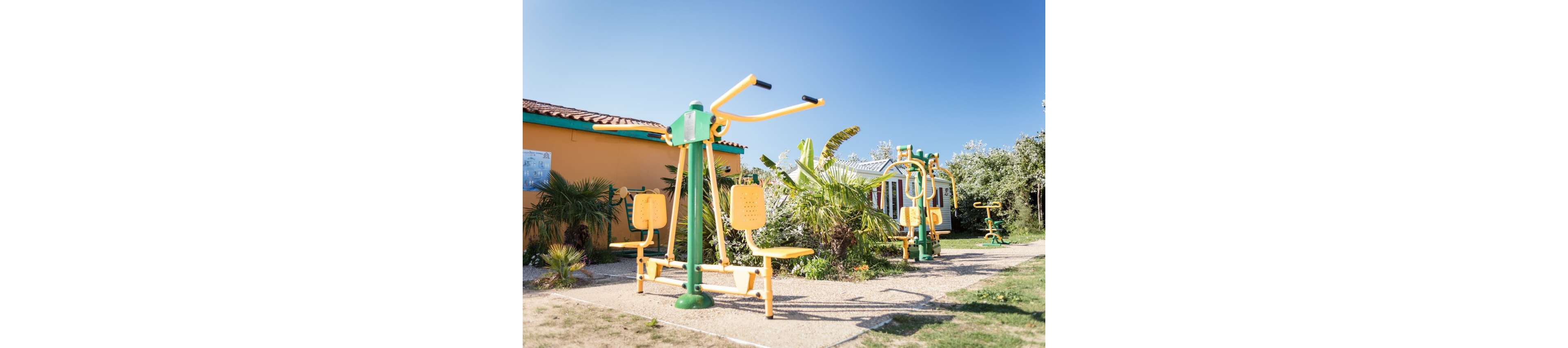 Bright yellow and green outdoor fitness machines stand along a gravel path in a sunny park. The equipment is placed near colorful low buildings and surrounded by palm trees and lush greenery. Clear blue skies and strong sunlight create a cheerful, inviting atmosphere. The scene suggests a recreational area designed for exercise and relaxation in a warm climate.