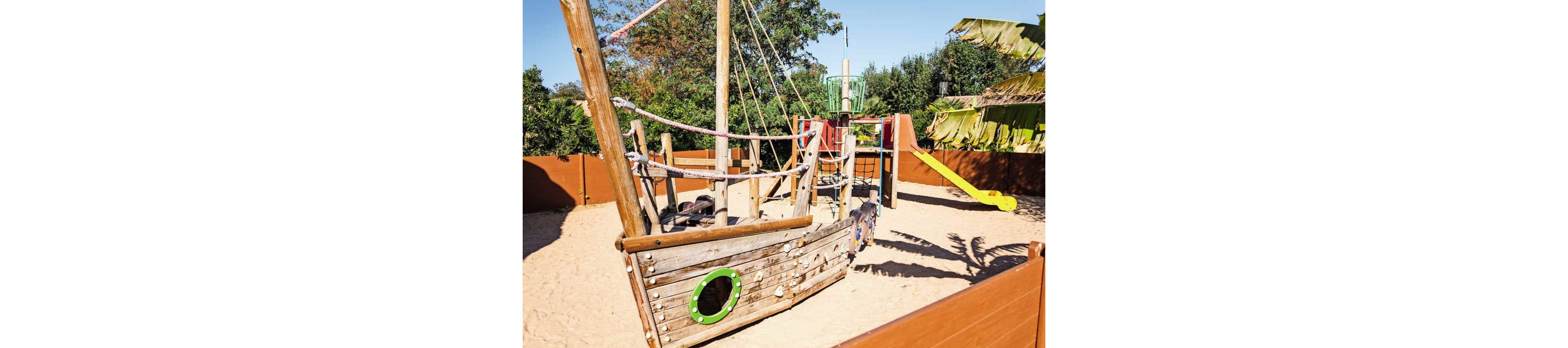 A wooden pirate ship themed playground structure stands in a large sandpit. The play area includes ropes, masts, and circular portholes on the ship. In the background, a slide and additional climbing elements are visible, surrounded by wooden fencing. Trees and greenery provide a natural outdoor setting under bright sunlight.