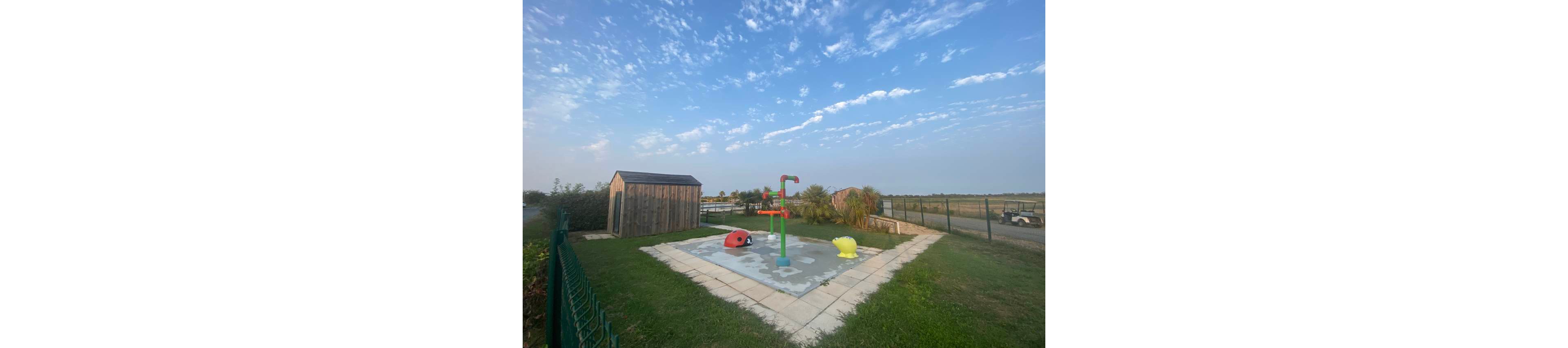 A small splash pad with colorful water play structures stands in a grassy outdoor area. A wooden shed is positioned nearby, and the pad is bordered by a tiled walkway and green fencing. The scene opens onto a rural landscape with open fields and a distant road. Soft morning or evening light and scattered clouds create a calm, relaxed atmosphere.