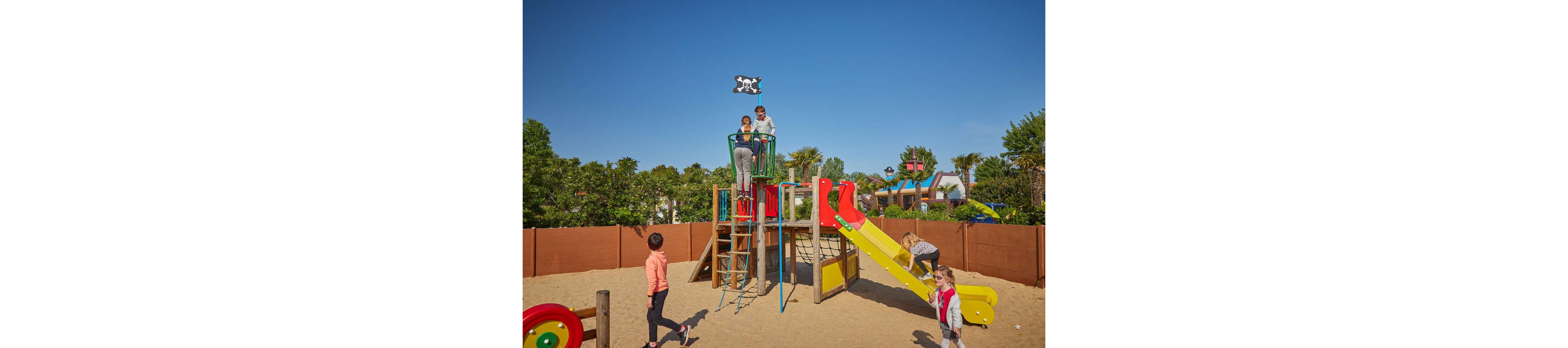 Several children play together on a colorful playground structure in a sandy outdoor play area. One child climbs a ladder while others stand on the platform beneath a small pirate flag. A bright yellow slide and red and blue elements add vibrant color to the scene. The playground is surrounded by a wooden fence with trees and greenery in the background under a clear blue sky.