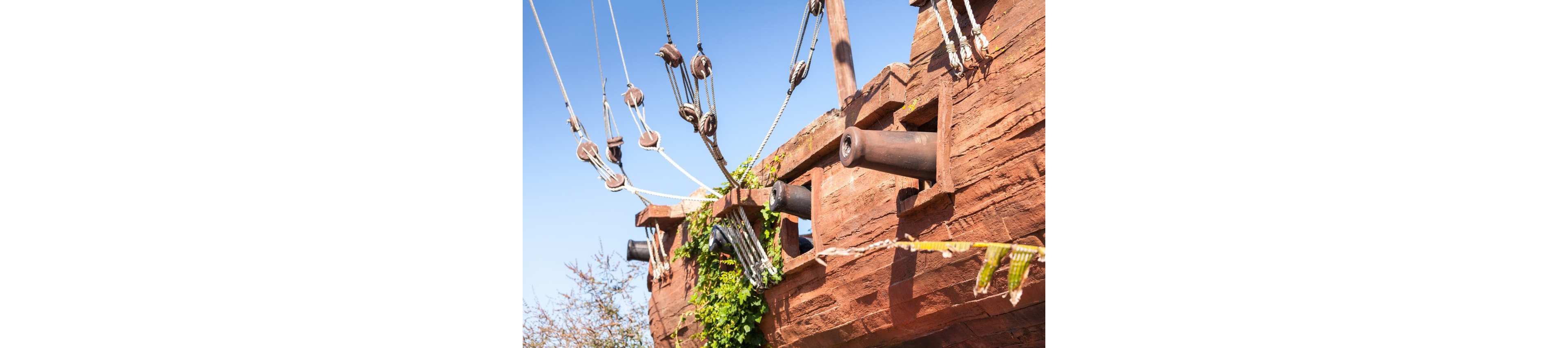 A rustic wooden ship structure features several cannons protruding from its side. Ropes, pulleys, and rigging hang from the masts against a clear blue sky. Green vines and foliage climb along the hull, suggesting age and weathering. The scene evokes a classic pirate or historical sailing ship atmosphere in bright daylight.