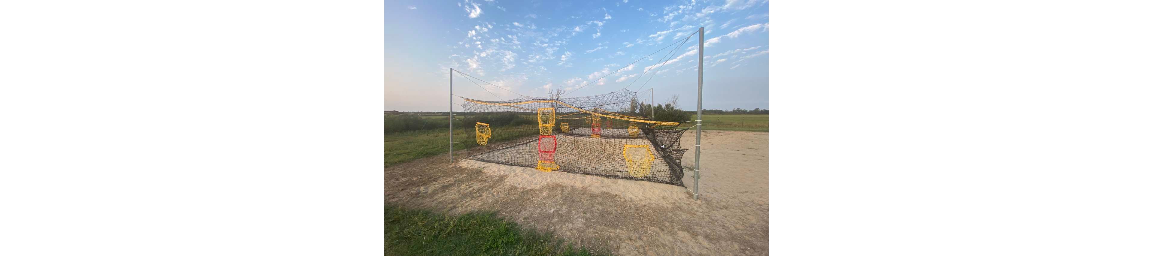 An outdoor practice field features a netted structure with multiple colorful target baskets suspended at different heights. The setup stands on a sandy or dirt surface surrounded by green grass and open countryside. Yellow and red plastic frames hang from the netting, creating a training or recreational target area. The sky above is partly cloudy with soft morning or evening light, giving the scene a calm rural atmosphere.