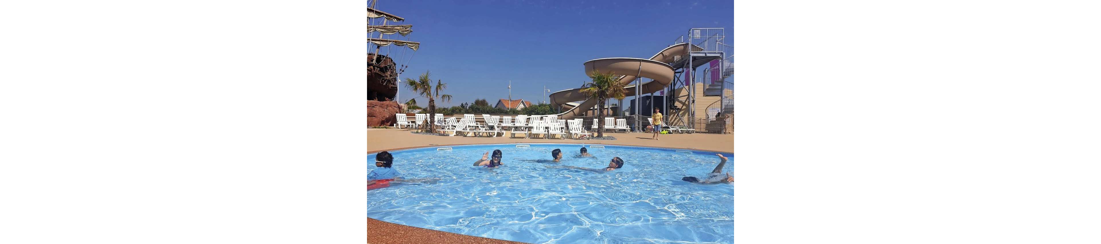 Several children swim and play in a shallow circular outdoor pool under a clear blue sky. In the background, white lounge chairs line the pool deck, and a large water slide structure rises to the right. A child in a yellow outfit walks near the slides, while palm trees and a ship-themed structure add to the resort atmosphere. The bright sunlight highlights the sparkling blue water and relaxed vacation setting.