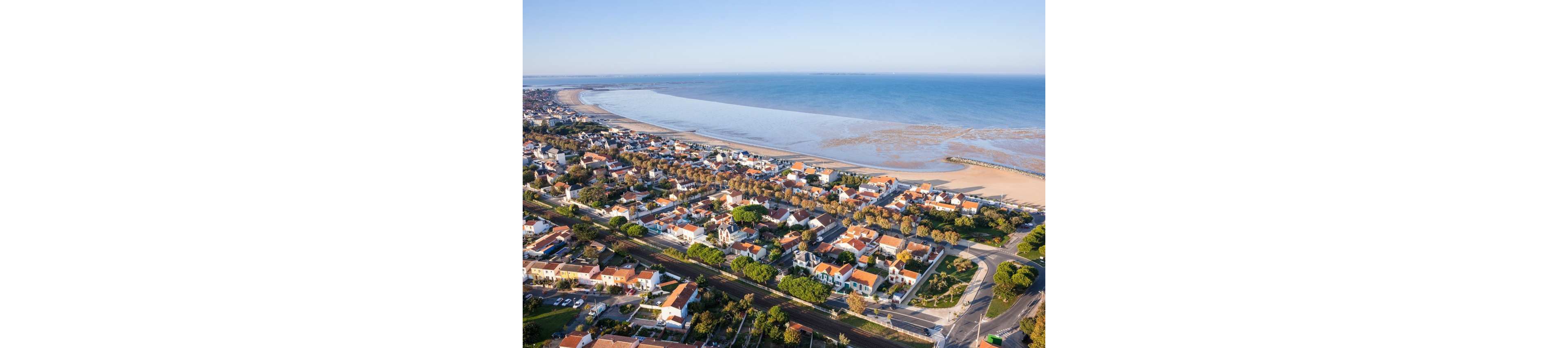 An aerial view shows a coastal town with rows of houses featuring red and white roofs near a wide sandy beach. The shoreline curves gently along the calm blue sea under clear skies. Streets, trees, and small buildings form an organized grid leading toward the waterfront. The overall scene feels peaceful and sunlit, highlighting a relaxed seaside community.
