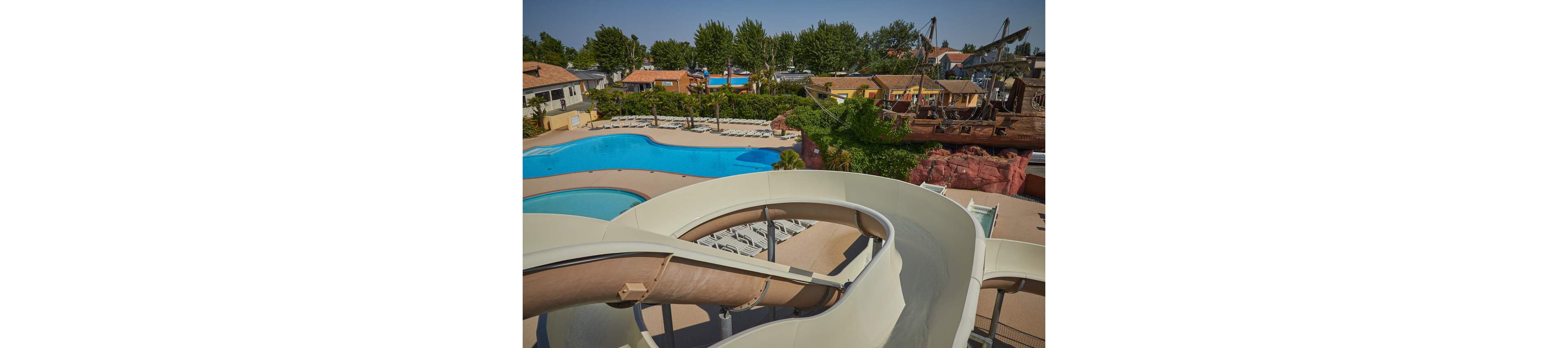 A large curving waterslide dominates the foreground of a resort-style water park. Below, a bright blue swimming pool is surrounded by rows of empty lounge chairs and sunbathing areas. In the background, there are trees, colorful buildings, and a themed pirate-ship structure adding visual interest. The scene appears calm and sunny, suggesting a leisure destination ready for visitors.